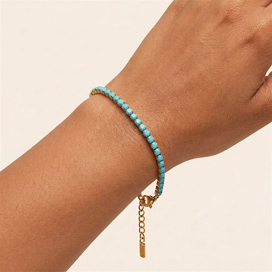 Close-up of a turquoise-blue CZ tennis bracelet worn on a wrist, with gold settings, a lobster clasp, and a hanging extender.