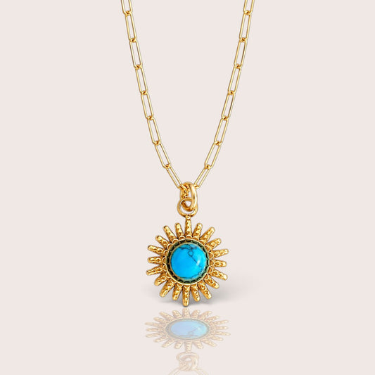 A pendant necklace featuring a sunburst design with a central round turquoise-blue gemstone, suspended from gold paperclip chain.
