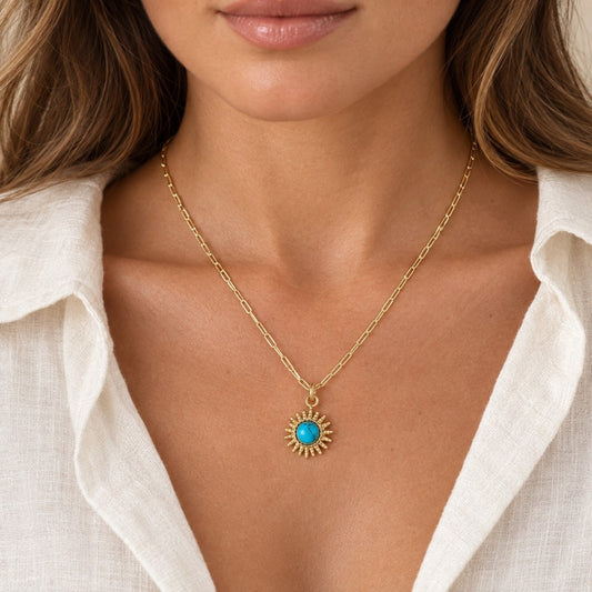 A mode with  gold chain necklace sunburst pendant featuring a round turquoise blue center, paired with an open white linen shirt.