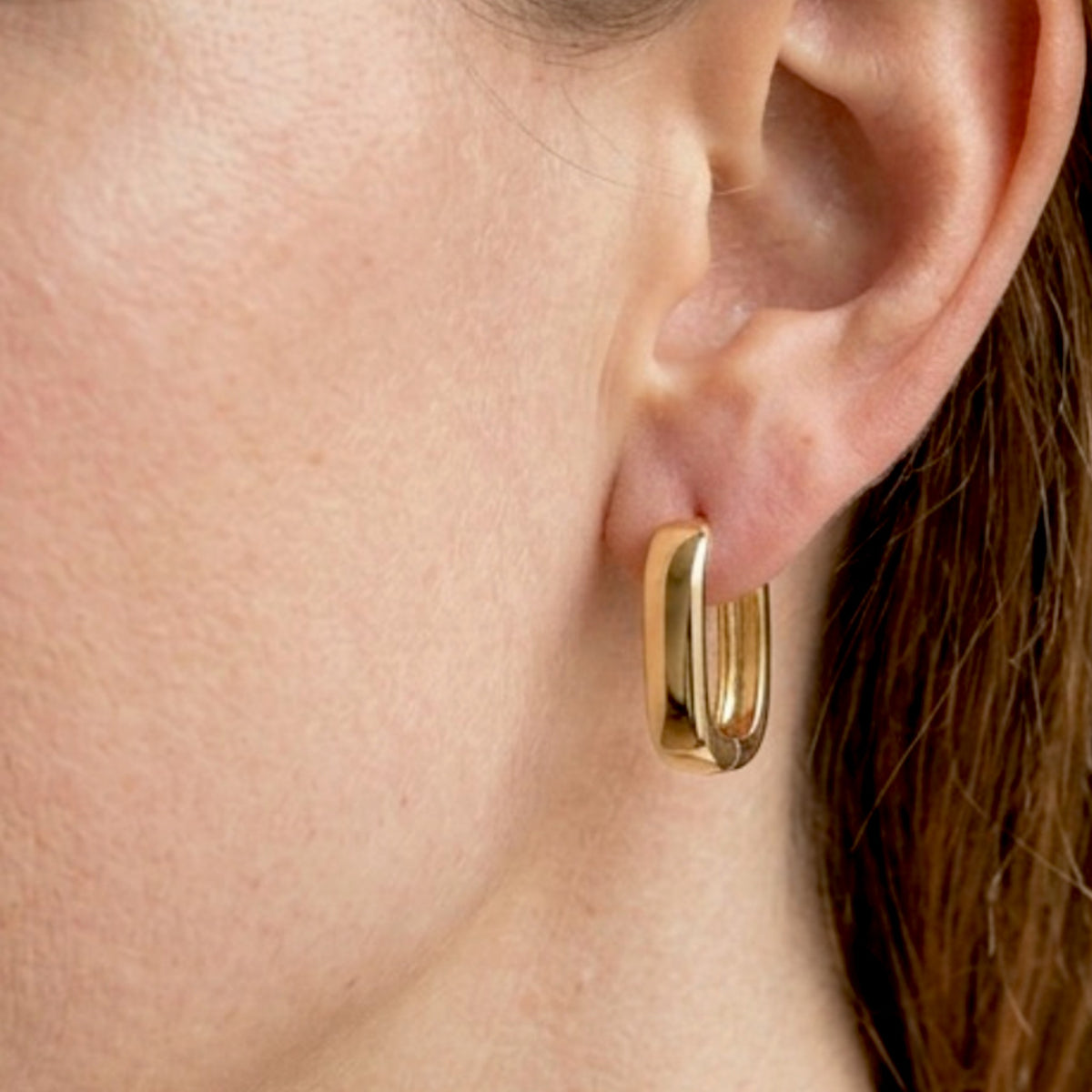 Close up of woman wearing a small gold rectangular huggie earring, with her hair tucked behind her ear.